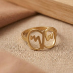 Gold ring with cut-out letter design on a beige fabric background